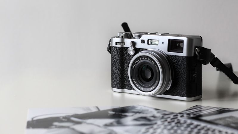 The image features a vintage-style camera placed on a flat surface alongside printed photographs. The camera is well-detailed, showcasing its lens and controls, while the photos appear to depict black-and-white imagery, suggesting a focus on photography as an art form.