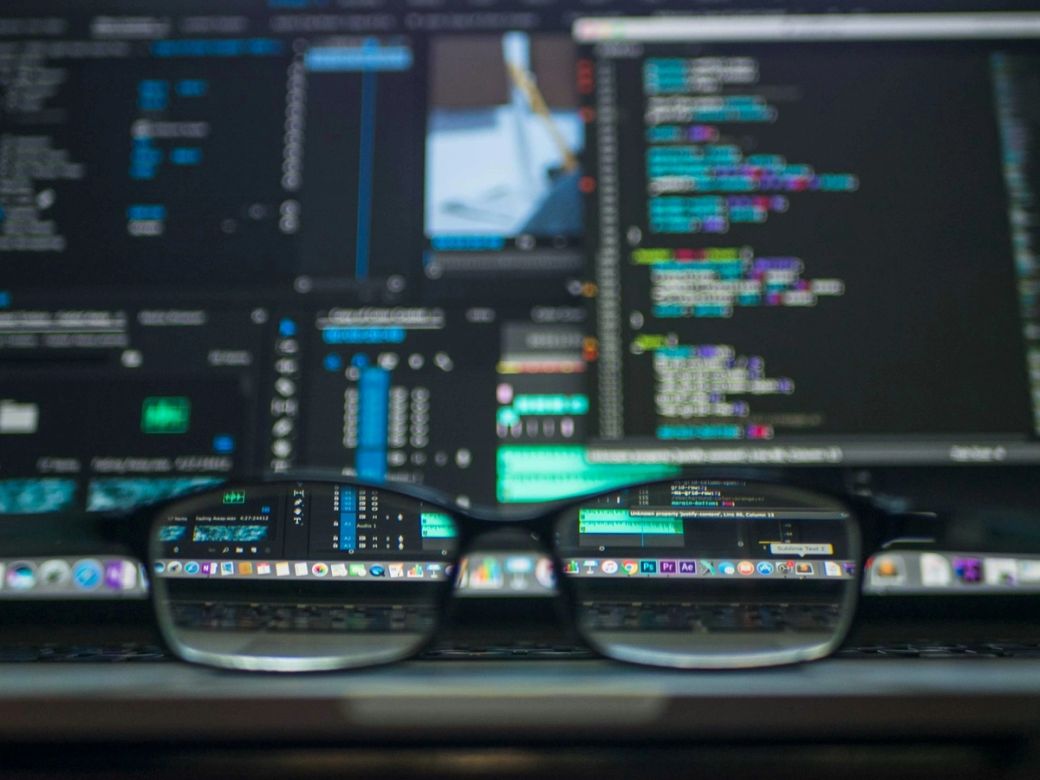 The image captures a close-up view of a pair of glasses in front of a computer screen displaying a variety of code and software interfaces. The reflections in the lenses show digital coding and graphical elements, suggesting a tech-related environment. — Credit: Photo by Kevin Ku / Unsplash