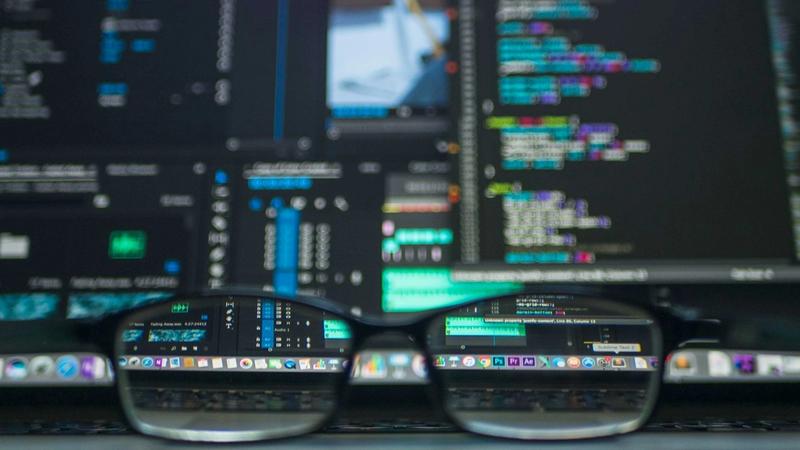 The image captures a close-up view of a pair of glasses in front of a computer screen displaying a variety of code and software interfaces. The reflections in the lenses show digital coding and graphical elements, suggesting a tech-related environment.