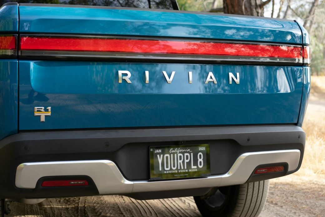 The image depicts the rear view of a blue Rivian electric vehicle parked on a dirt road surrounded by trees. The vehicle's distinctive taillights and license plate are clearly visible. — Credit: Photo by Clayton Cardinalli / Unsplash