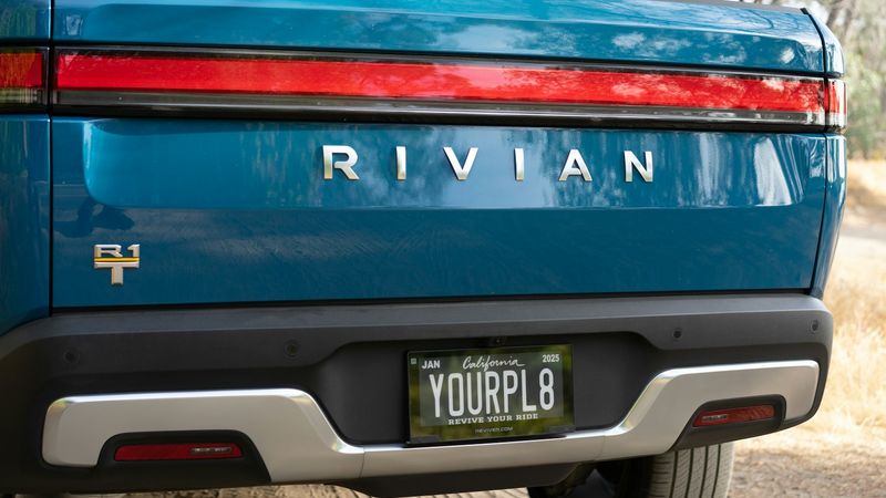 The image depicts the rear view of a blue Rivian electric vehicle parked on a dirt road surrounded by trees. The vehicle's distinctive taillights and license plate are clearly visible.