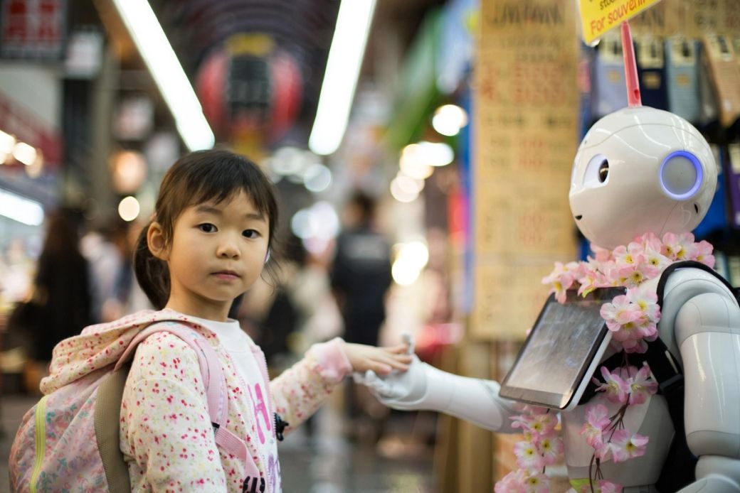 A child interacts with a humanoid robot in a busy market setting. The robot is decorated with flowers and has a screen display, while the background shows various market stalls. — Credit: Photo by Andy Kelly / Unsplash
