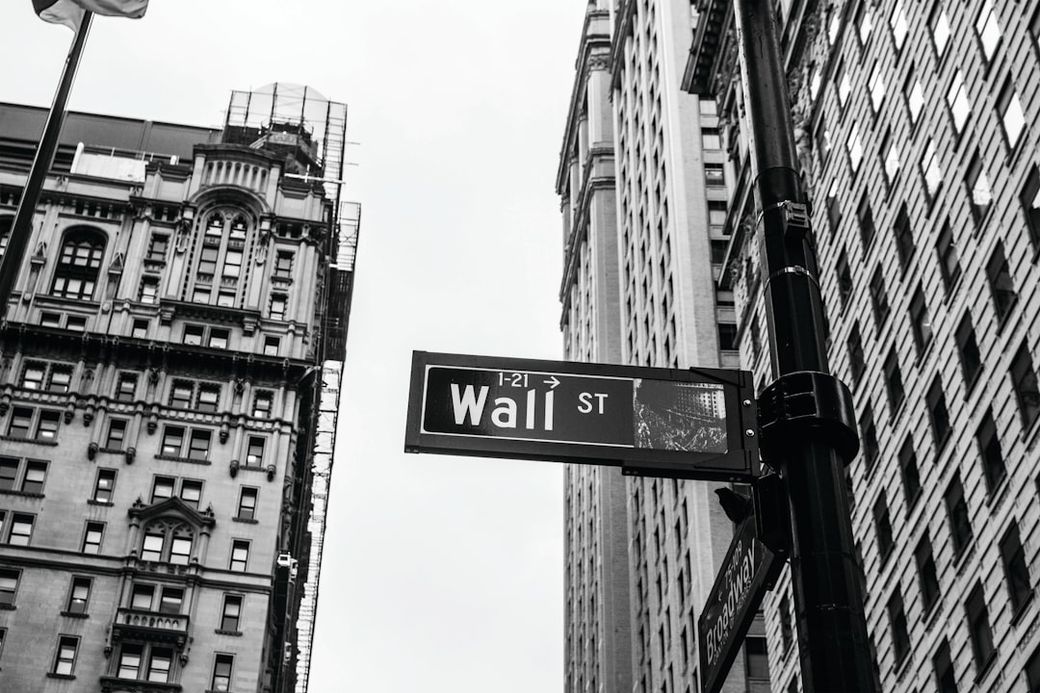 The image features a street sign for Wall Street, prominently displayed in a cityscape of tall buildings. The photograph is in black and white, emphasizing the architectural details of the surrounding structures. — Credit: Photo by Chris Li on Unsplash