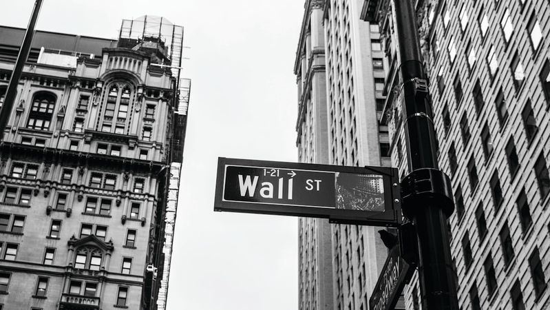 The image features a street sign for Wall Street, prominently displayed in a cityscape of tall buildings. The photograph is in black and white, emphasizing the architectural details of the surrounding structures.