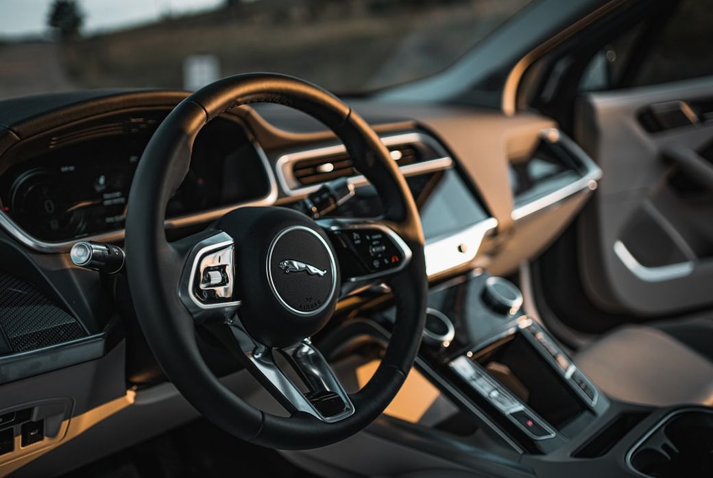 The interior of a luxury car featuring a modern dashboard layout and a circular steering wheel with a prominent logo. The dashboard includes digital displays and controls, showcasing a sleek design typical of high-end automobiles. — Credit: Photo by GRAHAM MANSFIELD on Unsplash