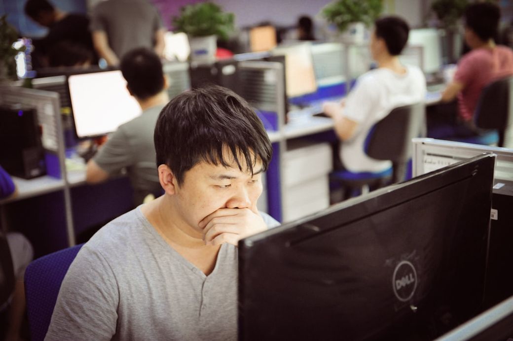 The image depicts an office environment with several individuals working at their computers. The focus is on a man in the foreground who appears to be deep in thought while looking at his screen. — Credit: Photo by blue sky on Unsplash