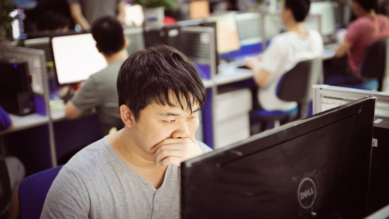 The image depicts an office environment with several individuals working at their computers. The focus is on a man in the foreground who appears to be deep in thought while looking at his screen.