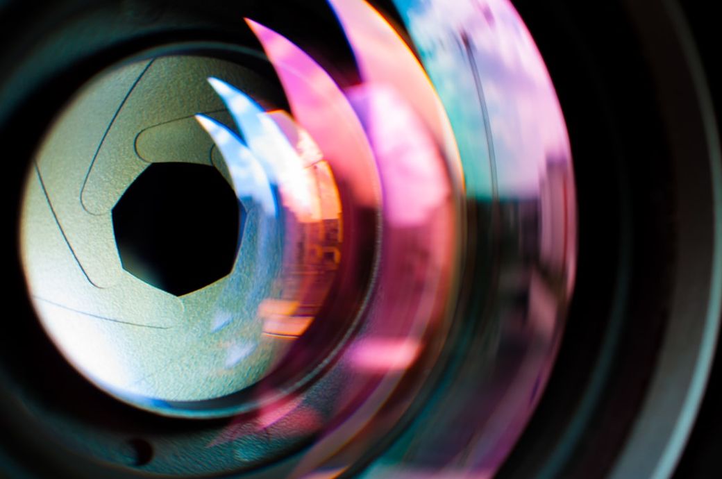 The image features an abstract close-up of a camera lens, showcasing the intricate details of its glass elements and reflections. The vibrant colors and shapes create a dynamic visual effect, emphasizing the technology of photography. — Credit: Photo by Agence Olloweb on Unsplash