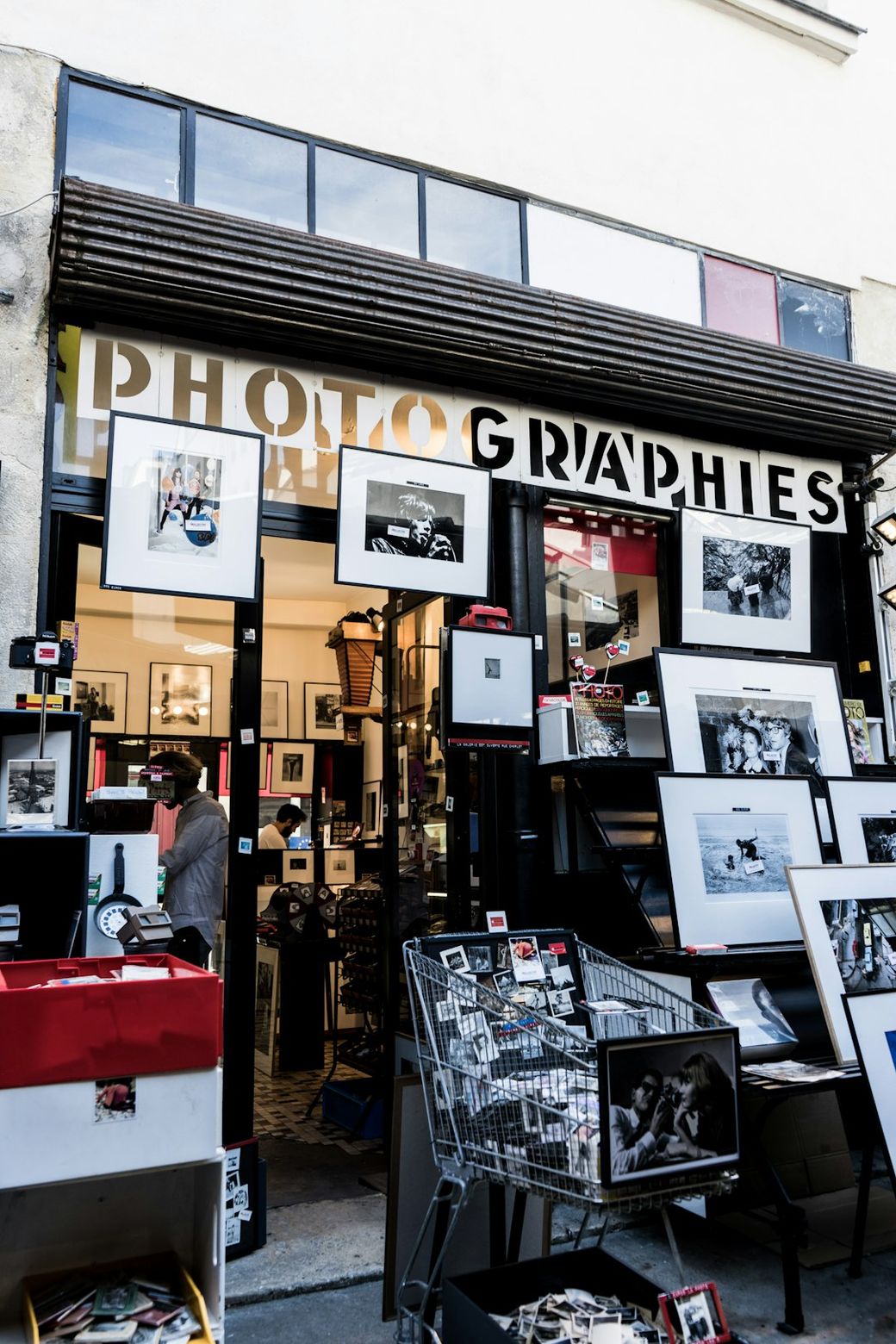 The image depicts the entrance of a photography shop, with various framed photographs displayed outside. The store features a sign that prominently reads 'PHOTOGRAPHIES' and has additional photographic artworks arranged in front. — Credit: Photo by Artiom Vallat on Unsplash