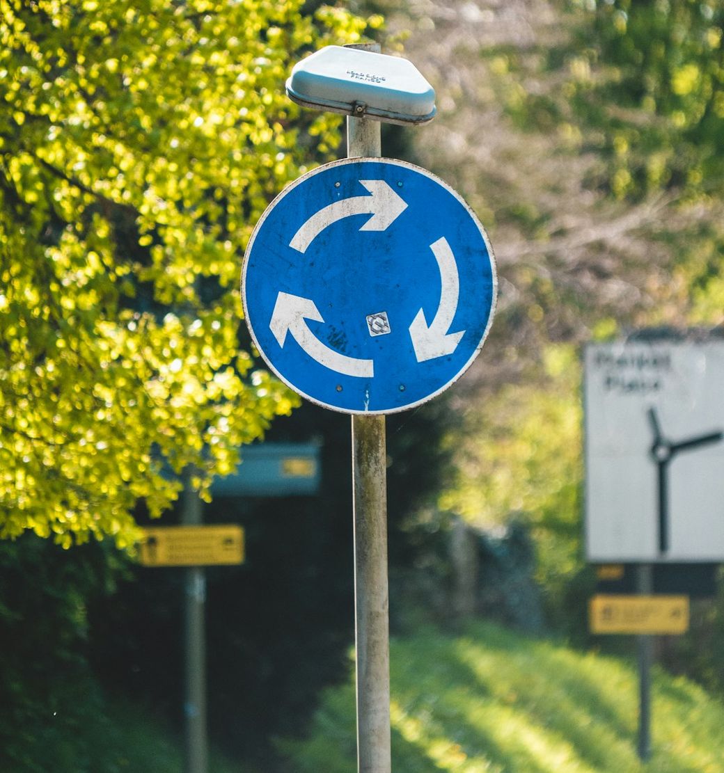 The image shows a circular road sign indicating a roundabout with four arrows pointing in a circular direction. In the background, there is blurred vegetation and additional signage partially visible. — Credit: Photo by Matt Seymour on Unsplash