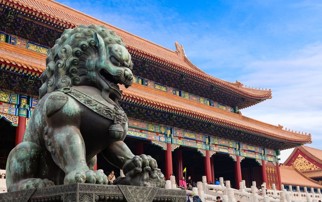 The image showcases a traditional architectural structure in a historical setting, highlighted by a prominent bronze lion statue in the foreground. The building features intricate detailing and vibrant colors characteristic of ancient Asian architecture. — Credit: Photo by Rafik Wahba on Unsplash