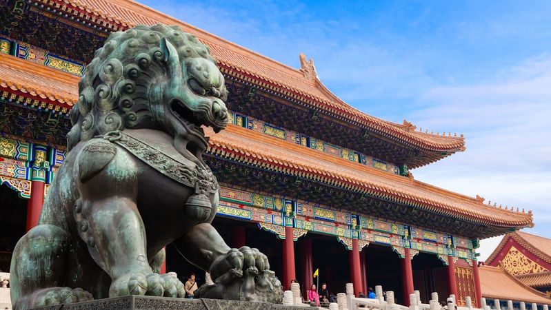 The image showcases a traditional architectural structure in a historical setting, highlighted by a prominent bronze lion statue in the foreground. The building features intricate detailing and vibrant colors characteristic of ancient Asian architecture.