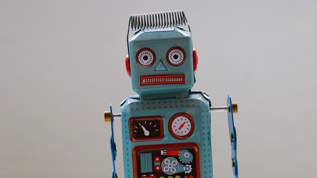 The image features a vintage toy robot with a retro design, displayed against a plain background. The robot has distinct facial features, including large eyes, and is adorned with mechanical details and dials. — Credit: Photo by Emilipothèse on Unsplash
