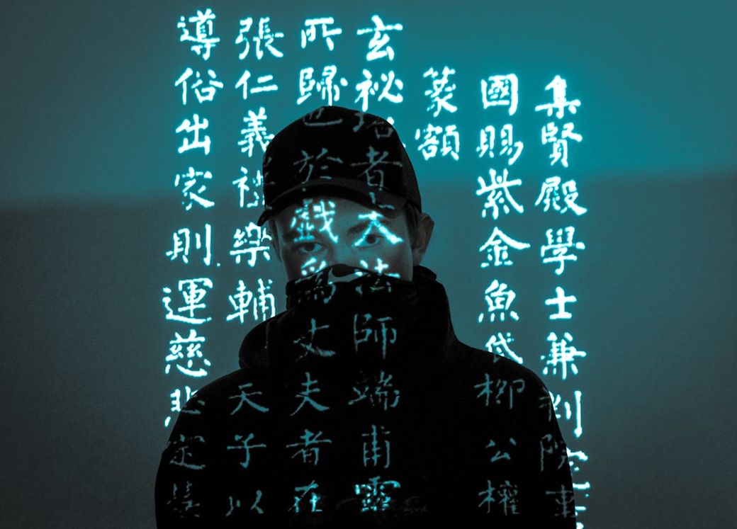 The image features a person wearing a cap and a mask, with projections of text in a foreign language illuminating their face. The background is a muted tone, enhancing the futuristic and mysterious atmosphere of the scene. — Credit: Photo by Pius Martin on Unsplash