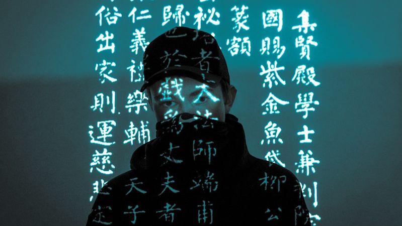 The image features a person wearing a cap and a mask, with projections of text in a foreign language illuminating their face. The background is a muted tone, enhancing the futuristic and mysterious atmosphere of the scene.