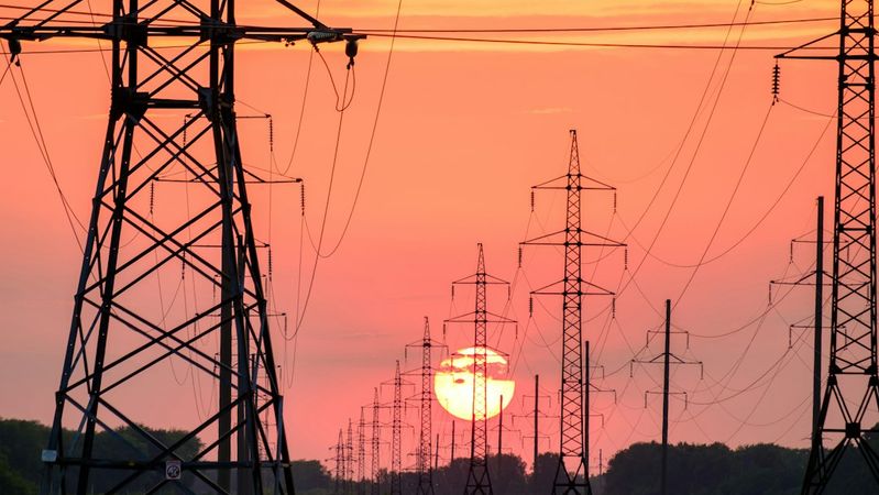The image features a landscape at sunset, dominated by several tall power transmission towers silhouetted against a colorful sky. The sun is setting on the horizon, creating a dramatic backdrop with orange and pink hues.