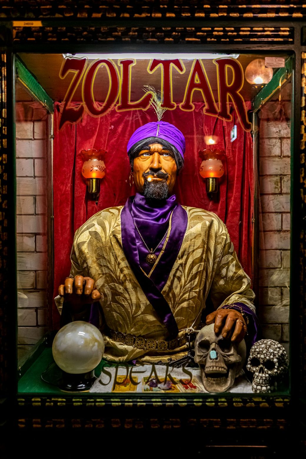 The image features a fortune-telling machine named Zoltar, depicting a stylized figure dressed in a purple turban and golden robe. The figure is positioned behind glass, surrounded by decorative skulls and a crystal ball, set against a backdrop of red velvet. — Credit: Photo by Hulki Okan Tabak on Unsplash
