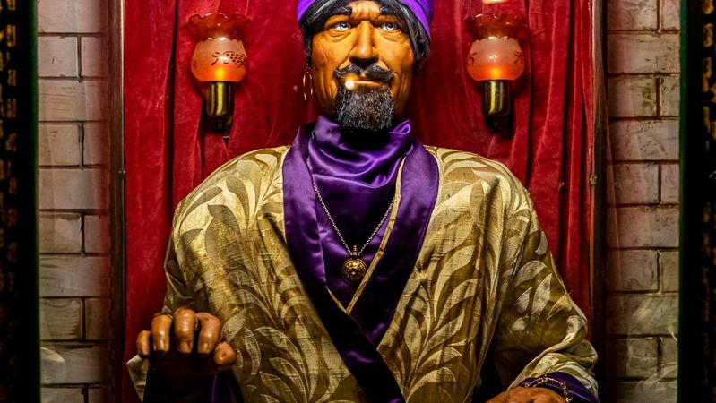 The image features a fortune-telling machine named Zoltar, depicting a stylized figure dressed in a purple turban and golden robe. The figure is positioned behind glass, surrounded by decorative skulls and a crystal ball, set against a backdrop of red velvet.
