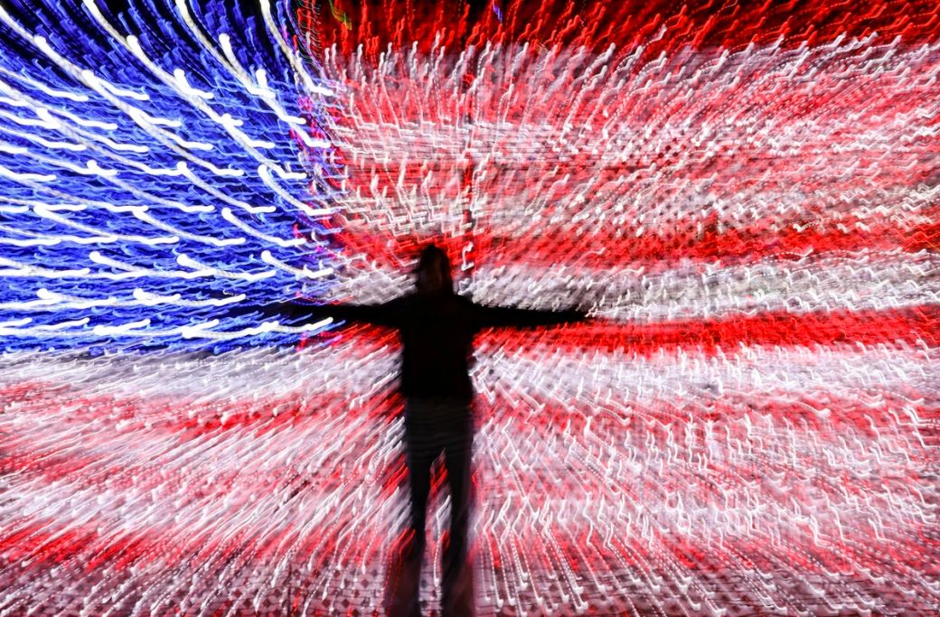 The image features a blurred figure standing against a vibrant backdrop designed to resemble the American flag, with swirling lights creating a dynamic effect. The contrast of the red, white, and blue colors conveys a sense of movement and energy. — Credit: Photo by Mick Haupt / Unsplash