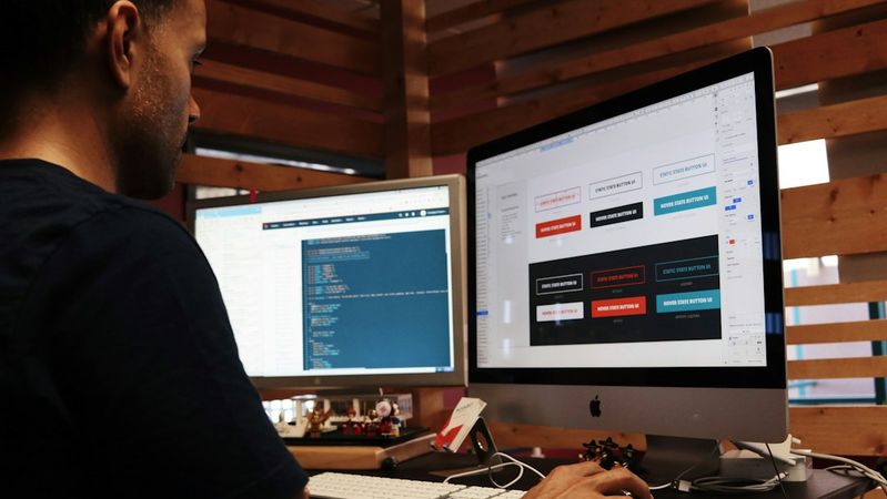 A man is seated at a desk working on a computer, focused on design elements displayed on the screen. The workspace features wooden paneling and multiple monitors, illustrating a modern office environment.