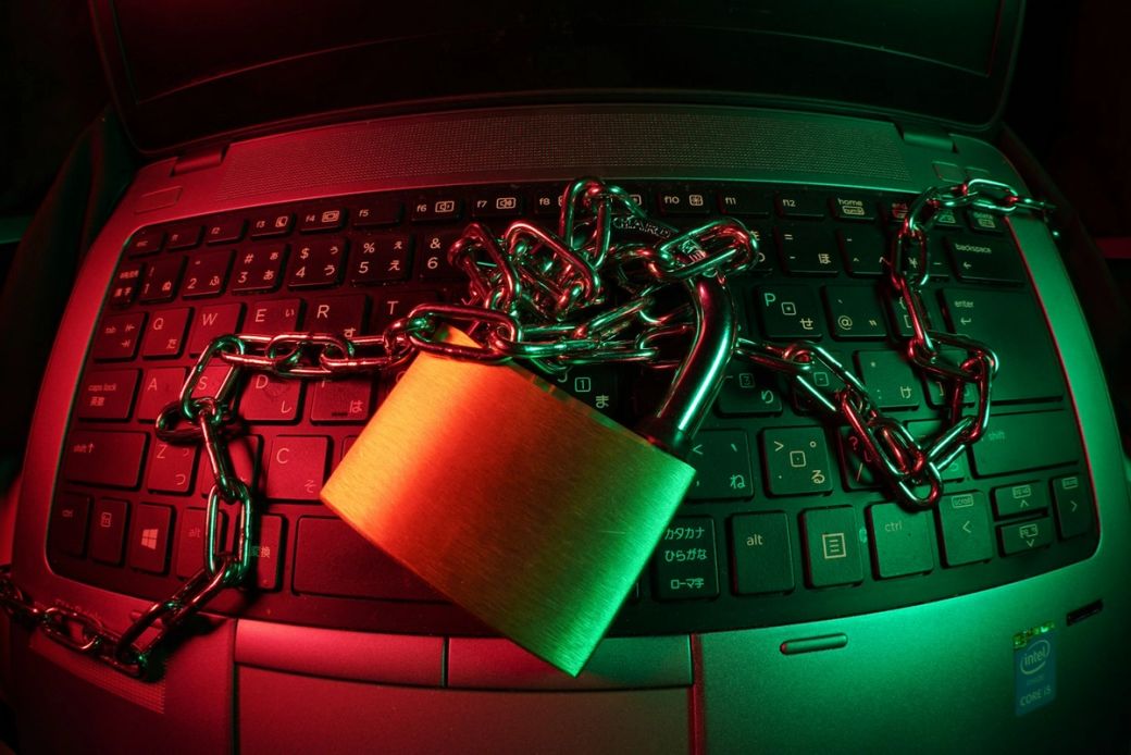 The image shows a close-up of a laptop keyboard with a large padlock and chains draped over it. The scene is illuminated with colorful lighting, highlighting the padlock and chains, suggesting themes of security and restriction. — Credit: Photo by FlyD / Unsplash