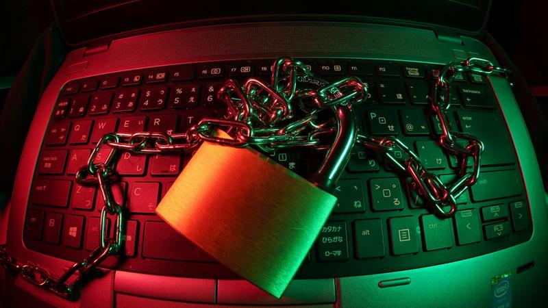 The image shows a close-up of a laptop keyboard with a large padlock and chains draped over it. The scene is illuminated with colorful lighting, highlighting the padlock and chains, suggesting themes of security and restriction.