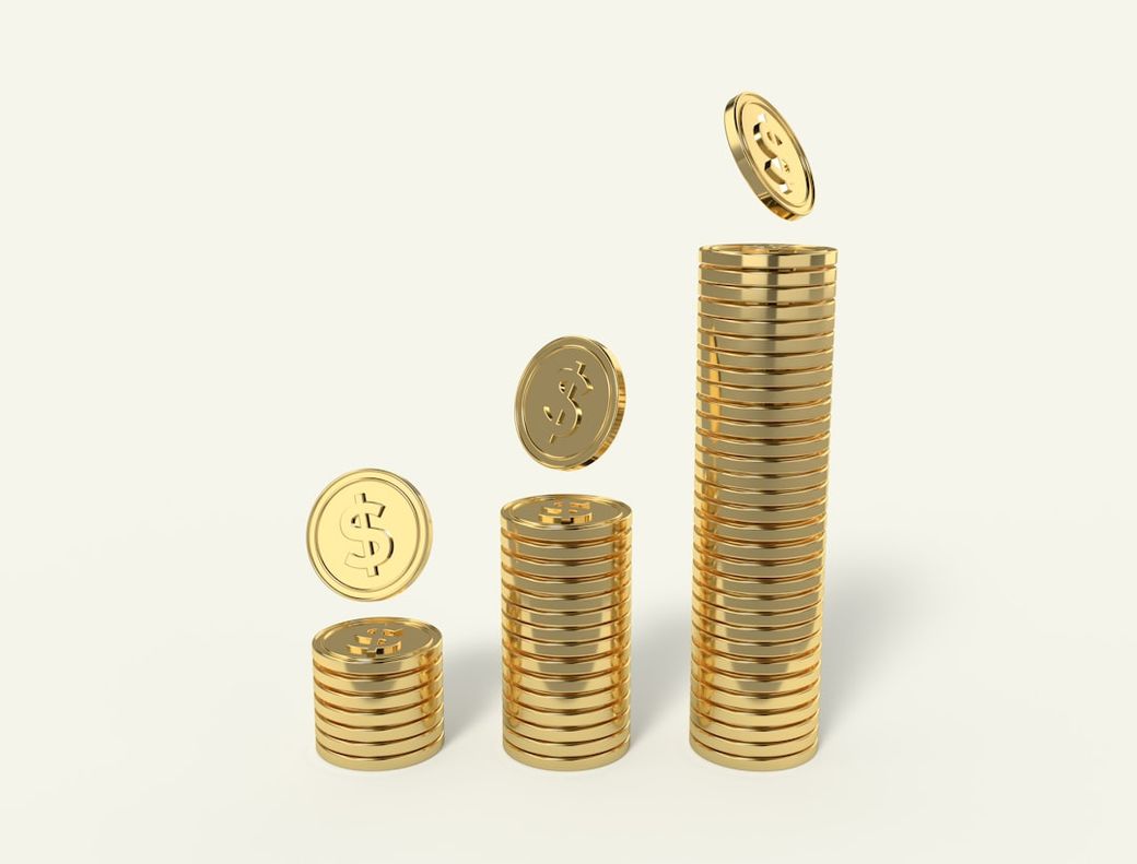 The image depicts golden coins stacked in ascending order, illustrating a concept of wealth accumulation and financial growth. Some coins are shown in mid-air as if they are being added to the stacks. — Credit: Photo by Allison Saeng on Unsplash