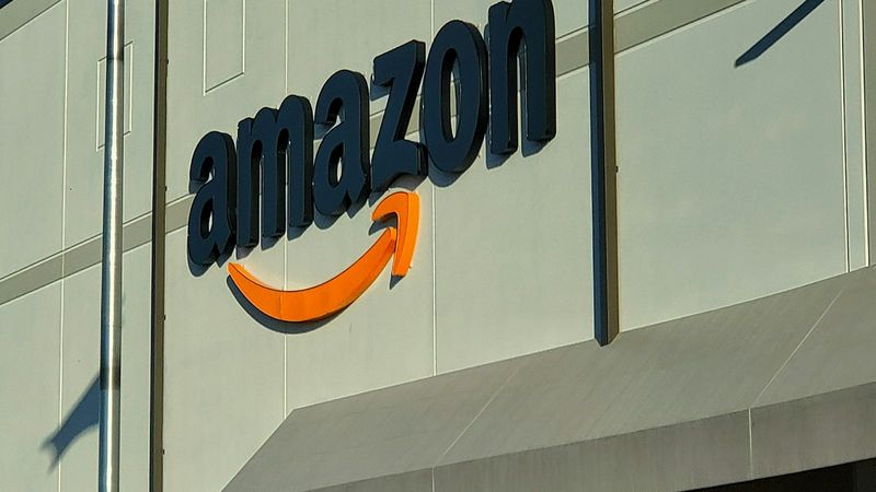 The image depicts the exterior of an Amazon facility, showcasing the company logo prominently above the entrance. A flag is seen waving in the breeze, and individuals are present near the entrance.