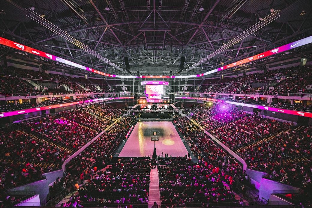 A large indoor arena filled with a diverse crowd attending an event. The brightly lit stage is prominent, surrounded by vibrant lighting and screens, creating an energetic atmosphere. — Credit: Photo by JC Gellidon on Unsplash