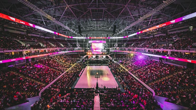 A large indoor arena filled with a diverse crowd attending an event. The brightly lit stage is prominent, surrounded by vibrant lighting and screens, creating an energetic atmosphere.