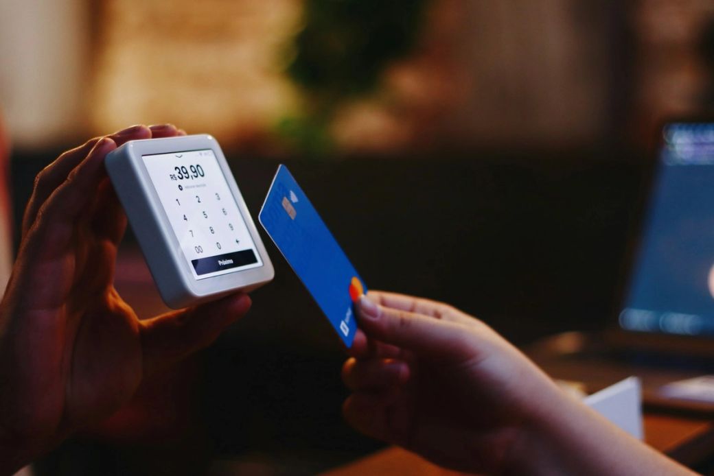 A person is holding a blue credit card in front of a payment terminal displaying a charge of $39.00. The background suggests a casual setting, possibly a cafe or restaurant. — Credit: Photo by Nathana Rebouças / Unsplash