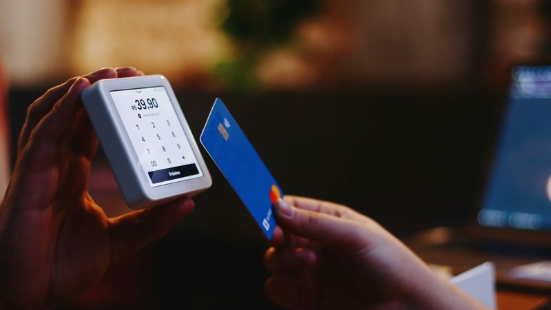 A person is holding a blue credit card in front of a payment terminal displaying a charge of $39.00. The background suggests a casual setting, possibly a cafe or restaurant.