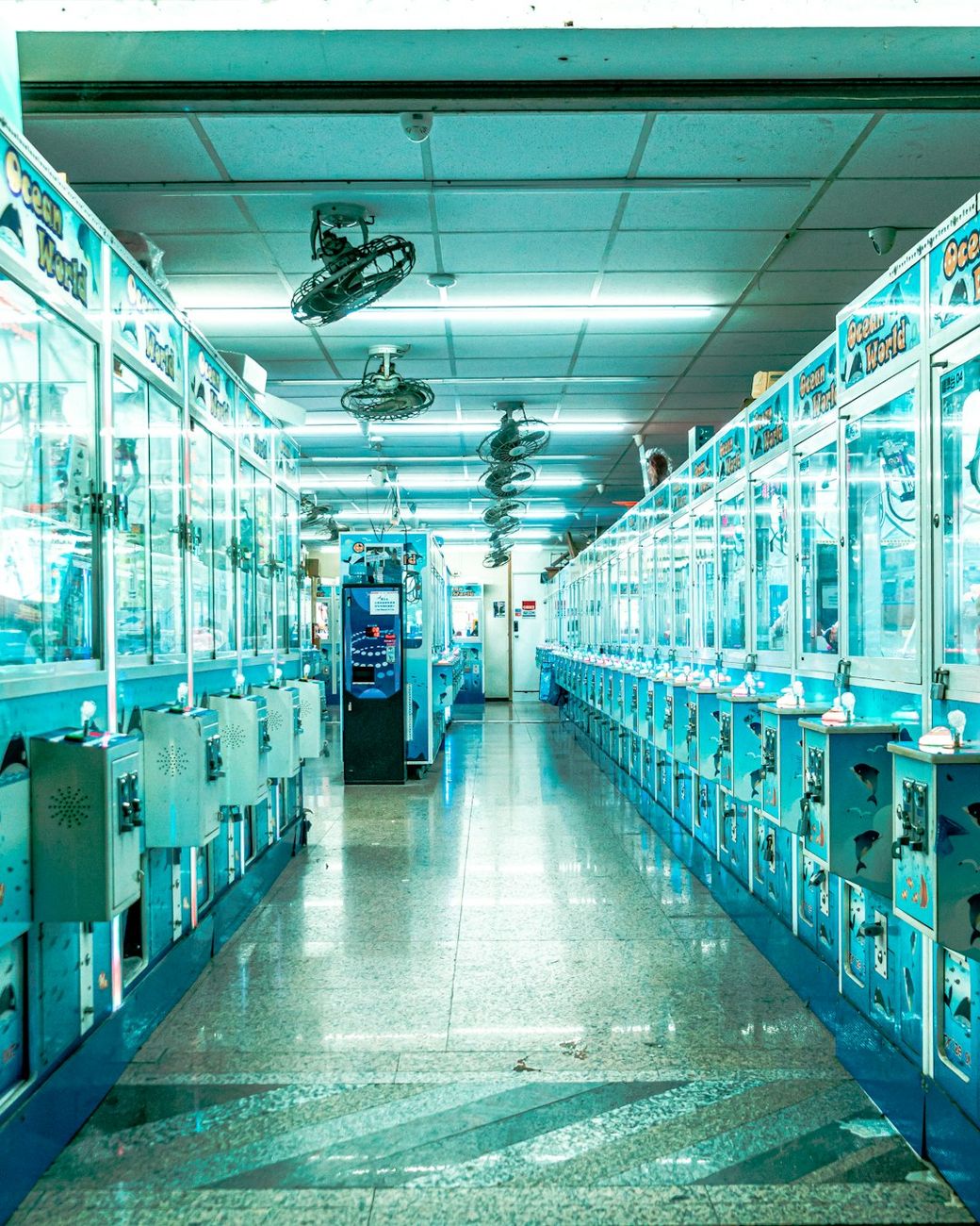 The image depicts a brightly lit indoor arcade filled with claw machines and vending kiosks. The perspective shows a long aisle that stretches towards a distant exit, with the machines lined up on either side. — Credit: Photo by Kai Cheng on Unsplash