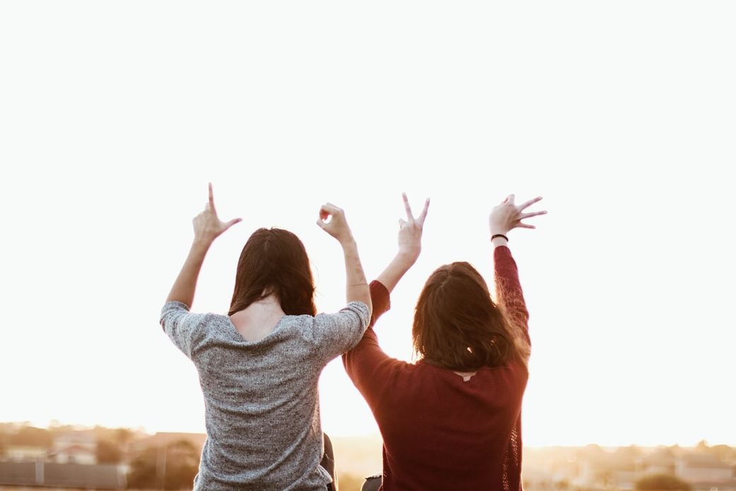 Two women stand with their backs to the camera, playfully gesturing towards the sky during sunset. Their poses suggest joy and celebration in a casual outdoor setting. — Credit: Photo by Sincerely Media on Unsplash