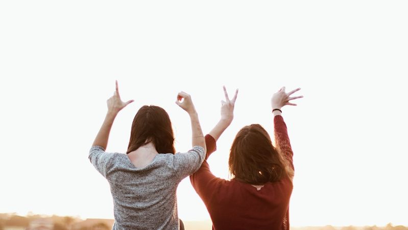 Two women stand with their backs to the camera, playfully gesturing towards the sky during sunset. Their poses suggest joy and celebration in a casual outdoor setting.