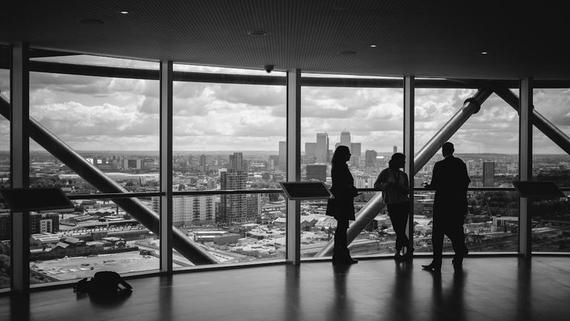 The image shows three silhouetted individuals engaged in conversation against a backdrop of a city skyline. The view from the large windows reveals a sprawling urban landscape under a cloudy sky.