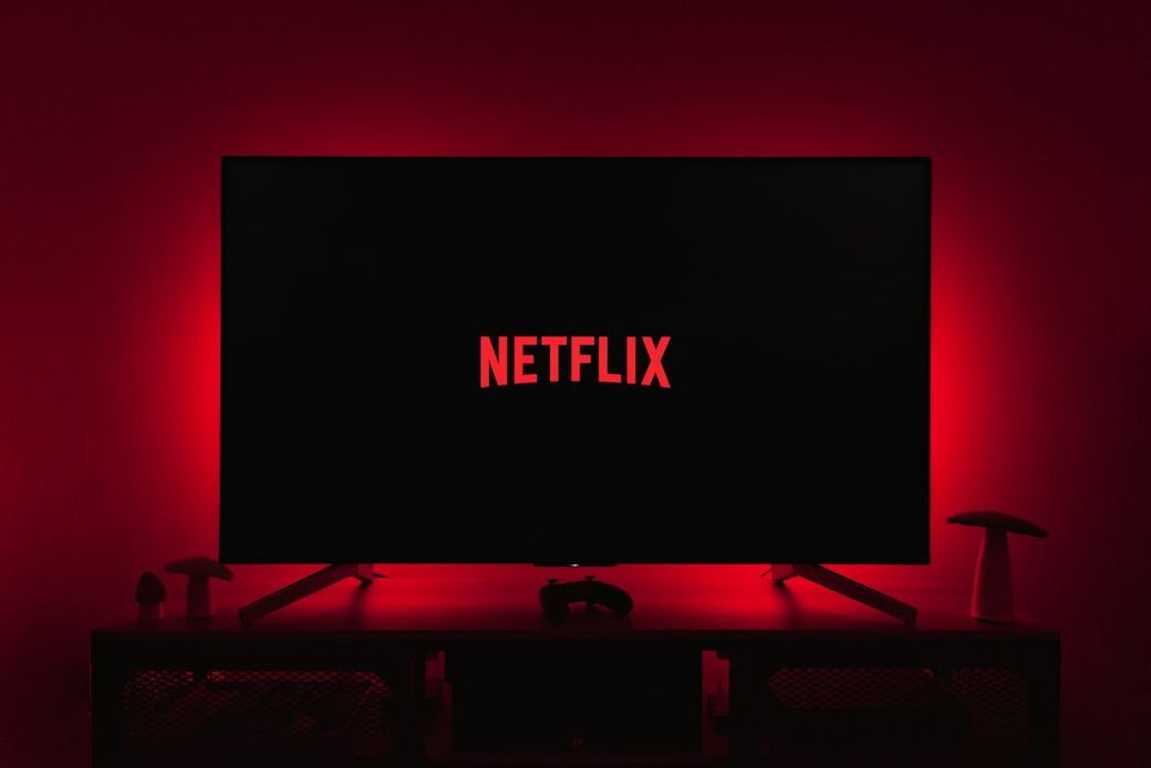 A television displaying the Netflix logo in a dimly lit room, highlighted by red ambient lighting. The setting includes a gaming console and decorative items on a shelf. — Credit: Photo by Thibault Penin on Unsplash