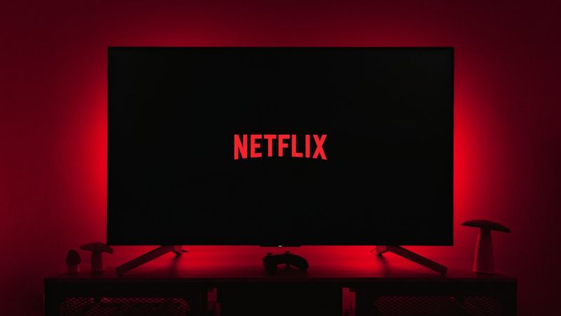 A television displaying the Netflix logo in a dimly lit room, highlighted by red ambient lighting. The setting includes a gaming console and decorative items on a shelf.