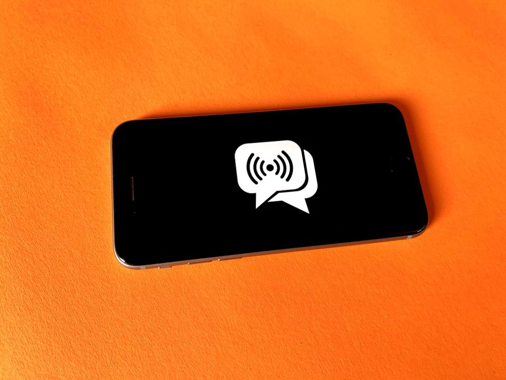The image features a smartphone laying on a vibrant orange surface. The screen displays a speech bubble icon with sound waves, suggesting communication or audio transmission. — Credit: Photo by Franck on Unsplash