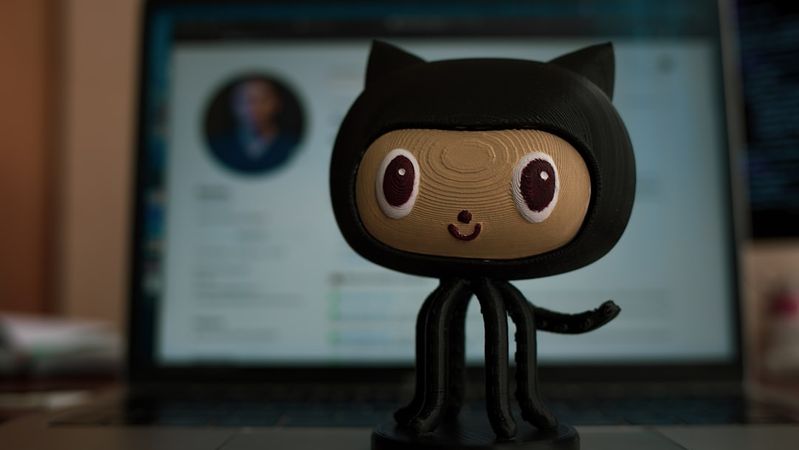 The image features a 3D-printed character resembling a cat with large eyes, positioned in the foreground. In the background, there is a laptop displaying a user's profile on a digital platform, hinting at a tech-related context.