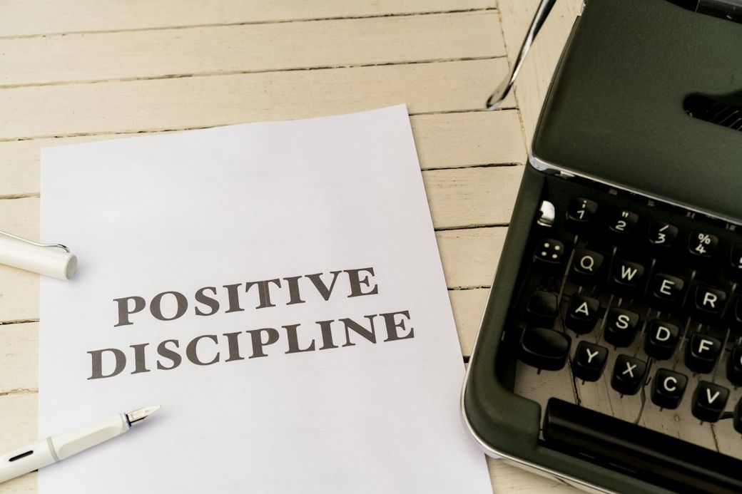 The image shows a printed sheet of paper with the words 'POSITIVE DISCIPLINE' prominently displayed. It is situated on a wooden surface next to a vintage typewriter and a pen. — Credit: Photo by Markus Winkler on Unsplash