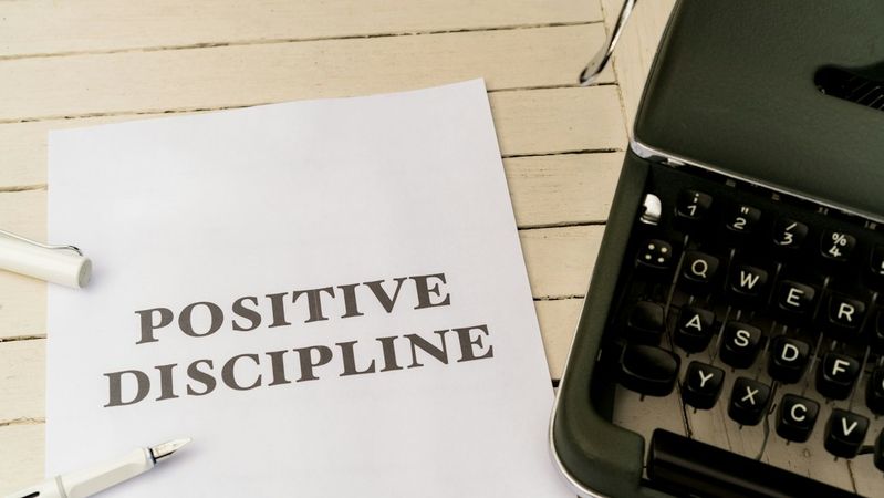 The image shows a printed sheet of paper with the words 'POSITIVE DISCIPLINE' prominently displayed. It is situated on a wooden surface next to a vintage typewriter and a pen.
