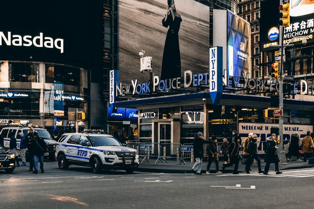 The image captures a bustling scene in Times Square, New York City, featuring the New York Police Department (NYPD) precinct. Passersby walk across the intersection, with several police vehicles visible. — Credit: Photo by Meriç Dağlı on Unsplash