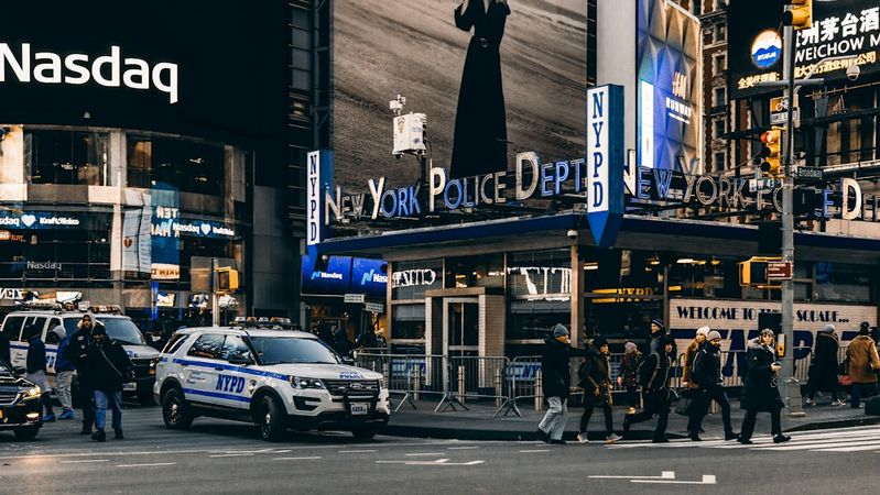 The image captures a bustling scene in Times Square, New York City, featuring the New York Police Department (NYPD) precinct. Passersby walk across the intersection, with several police vehicles visible.