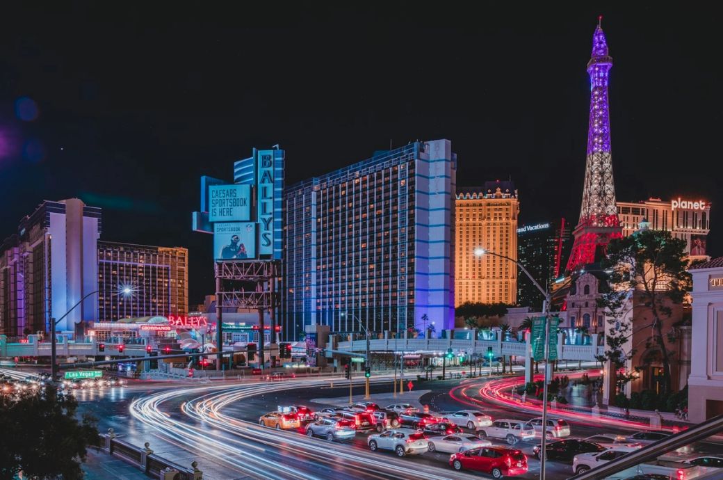 A bustling night scene of the Las Vegas Strip, highlighting prominent buildings and landmarks illuminated by colorful lights. The Eiffel Tower replica and Bally's hotel stand out amid the vibrant city atmosphere. — Credit: Photo by Paul C Barranco / Unsplash