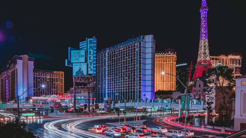 A bustling night scene of the Las Vegas Strip, highlighting prominent buildings and landmarks illuminated by colorful lights. The Eiffel Tower replica and Bally's hotel stand out amid the vibrant city atmosphere.