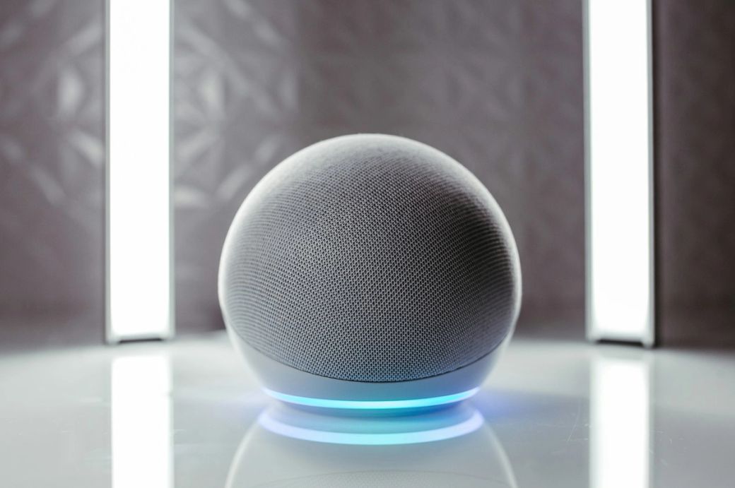 This image features a smart speaker with a spherical design, positioned on a reflective surface. The background includes two vertical light sources, creating a modern and sleek atmosphere. — Credit: Photo by Jonathan Borba / Unsplash