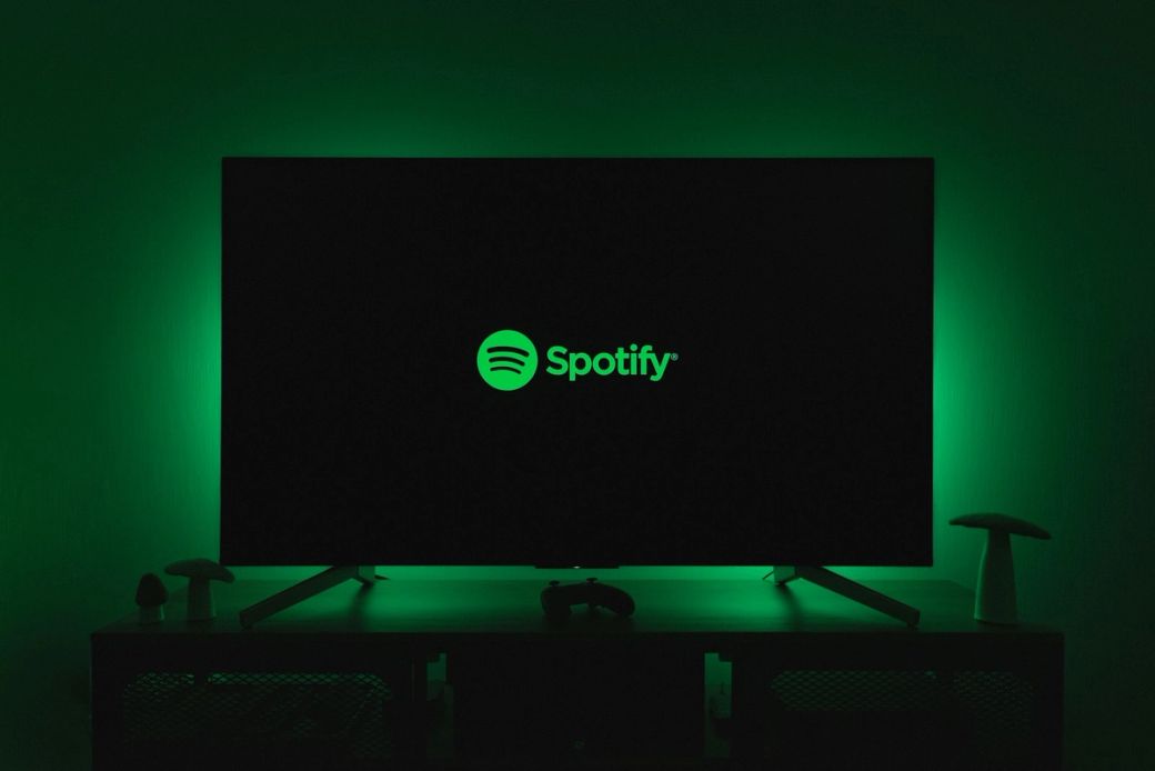 An illuminated television displaying the Spotify logo in a dark room. The ambiance is enhanced by a green backlight, creating a modern digital environment. — Credit: Photo by Thibault Penin / Unsplash