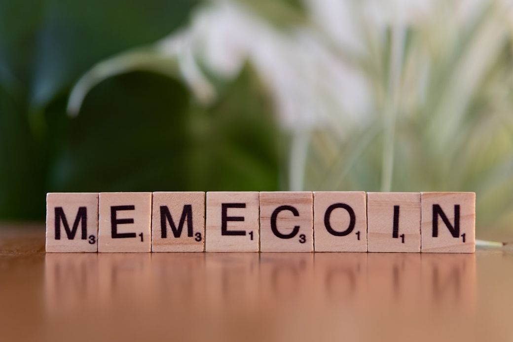 The image features wooden blocks displaying the word 'MEMECOIN'. The background has a soft, blurred green appearance, suggesting a casual or playful setting. — Credit: Photo by Markus Winkler on Unsplash