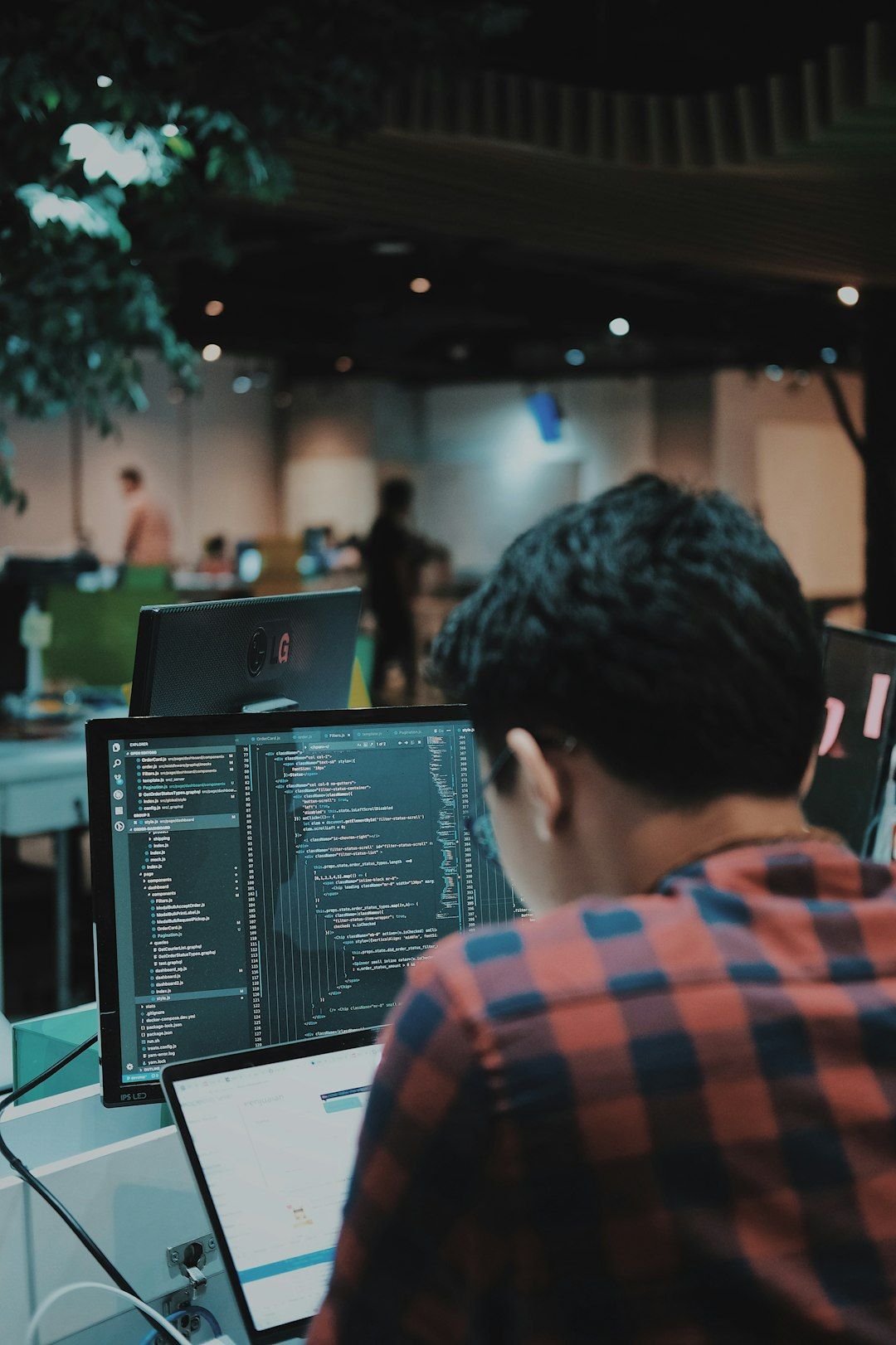 A man is seated at a computer desk, focused on programming on his laptop while multiple screens display code. The workspace has a modern design with greenery in the background and other individuals working in a collaborative environment.